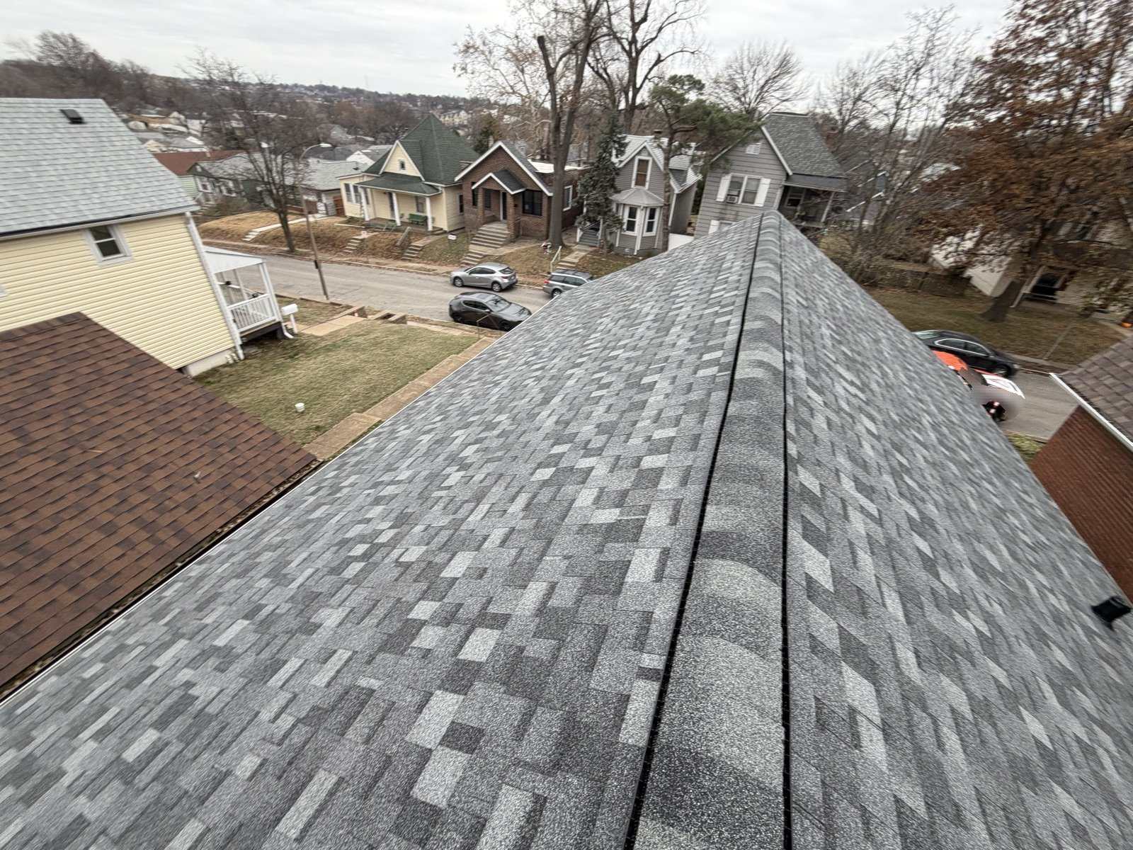 Grey architectural shingles on a finished roof photographed along the ridge, with a continuous ridge vent running the length and an older street of brick and frame St. Louis houses below