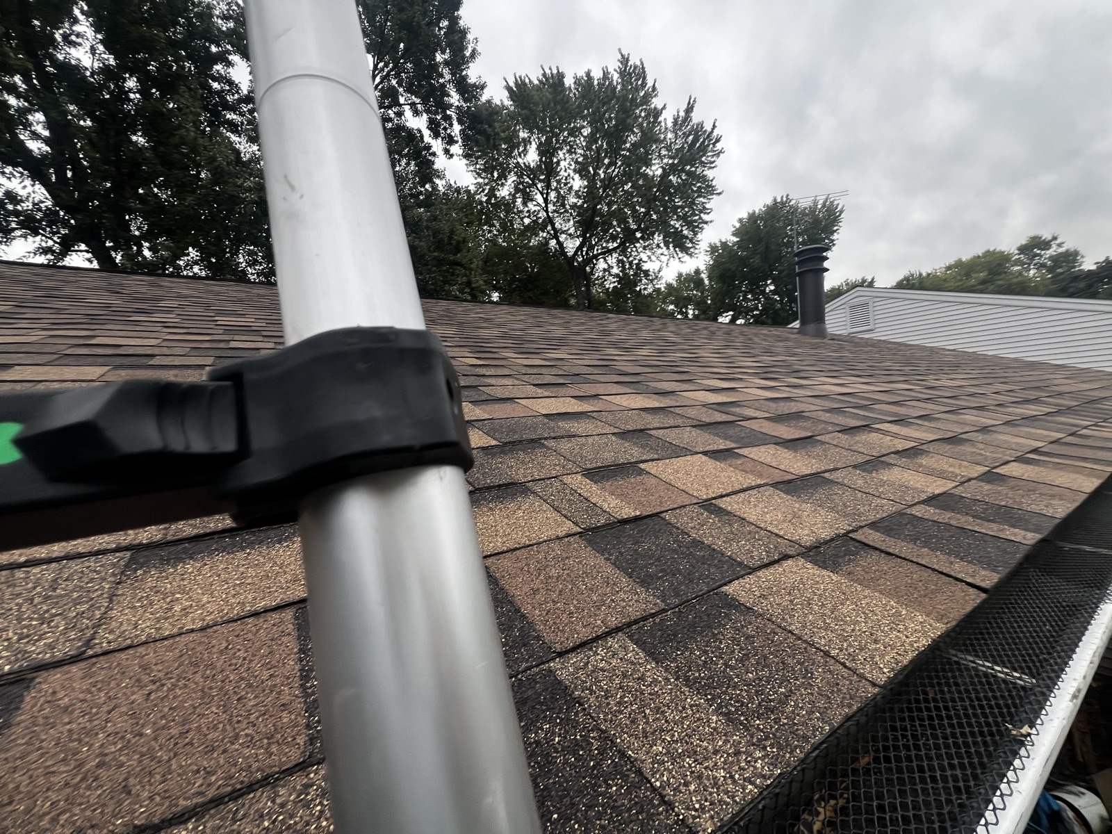 A ladder stabiliser resting on a weathered brown shingle roof, with a mesh gutter guard running along the eave