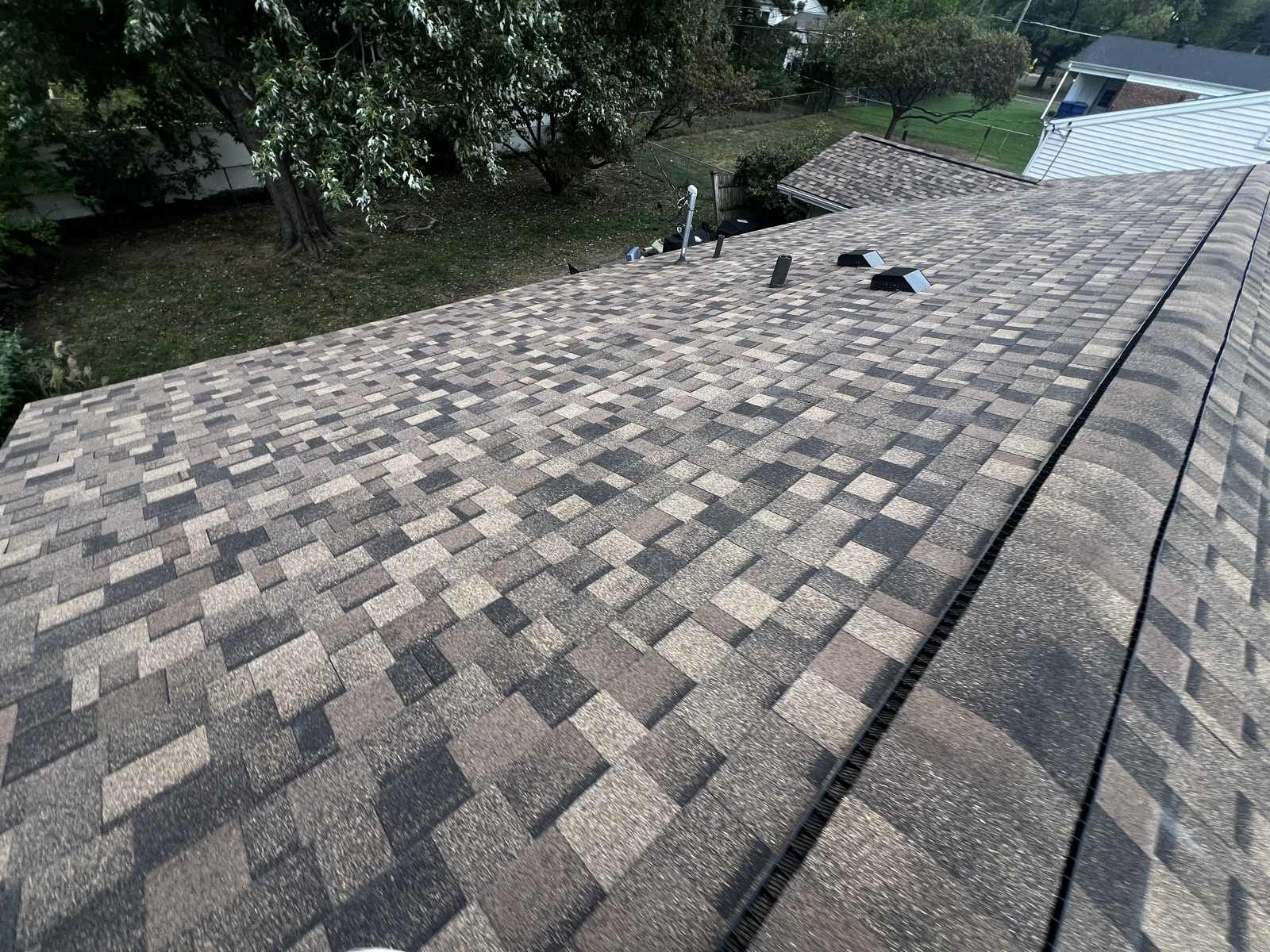 Broad expanse of a newly laid brown, tan and charcoal multi-tone architectural shingle roof, with the capped ridge and a dark ridge vent running down one side and two black box vents near the far end
