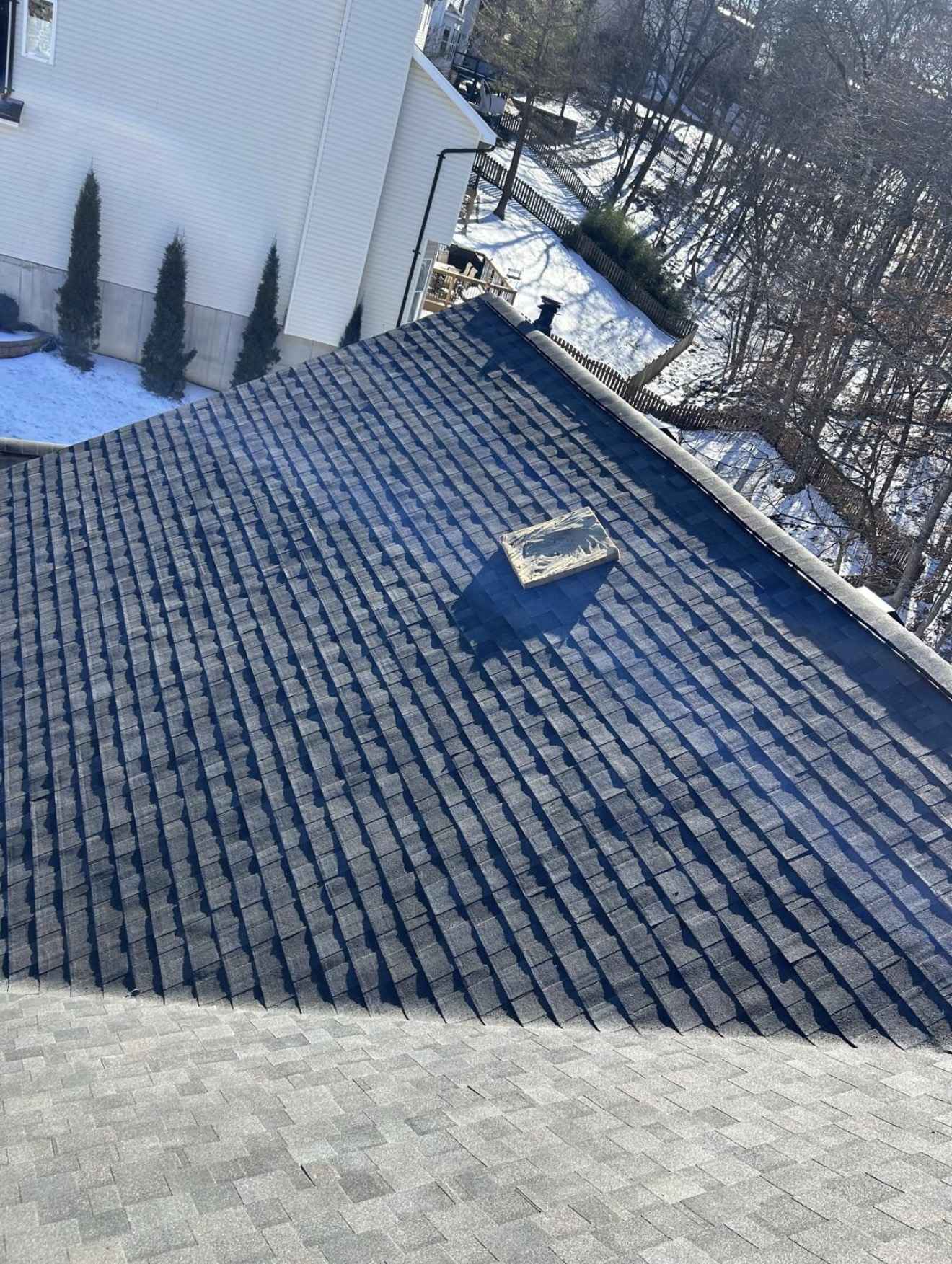 Part laid charcoal architectural shingles running down a roof plane in winter, meeting a lighter grey shingle surface across the foreground, with an offcut lying loose on the slope and snow on the ground beyond
