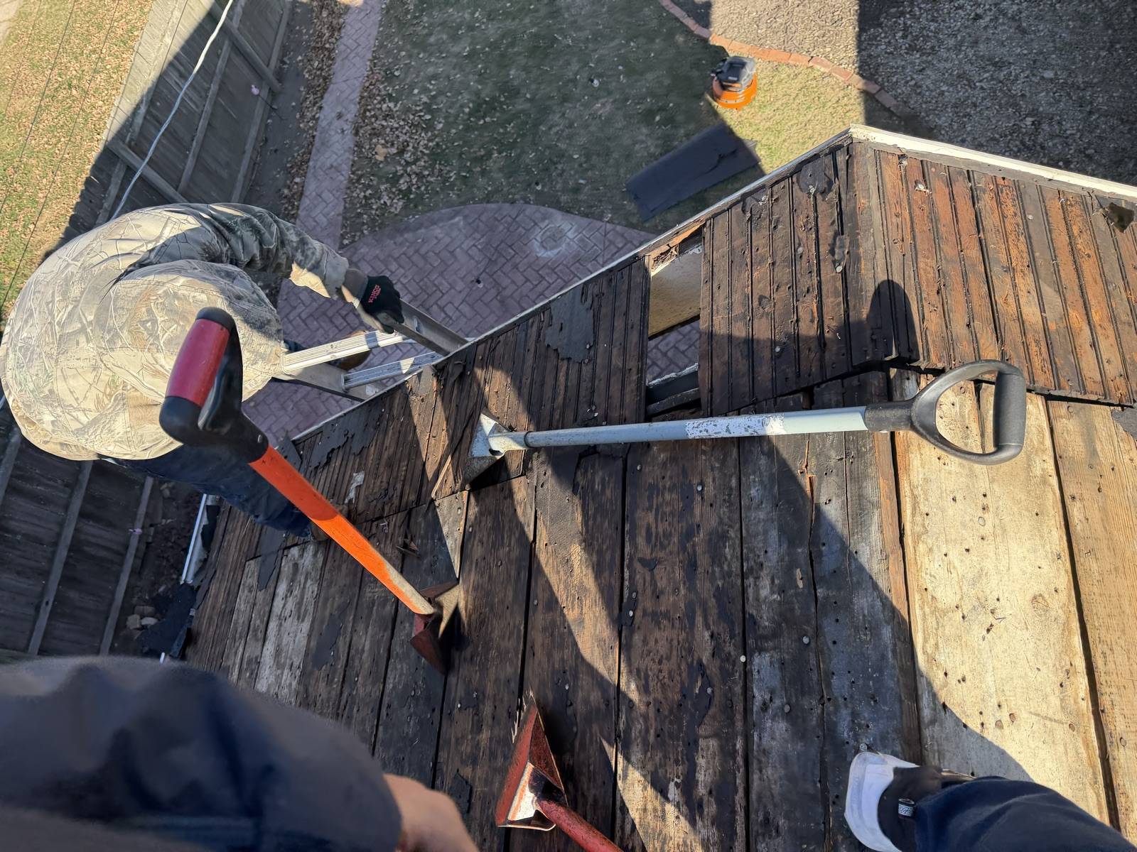 Tear-off bars, a roofing shovel and two flat spades lying on stripped plank roof decking, with an aluminium ladder hooked over the eave and a worker crouched at the far edge