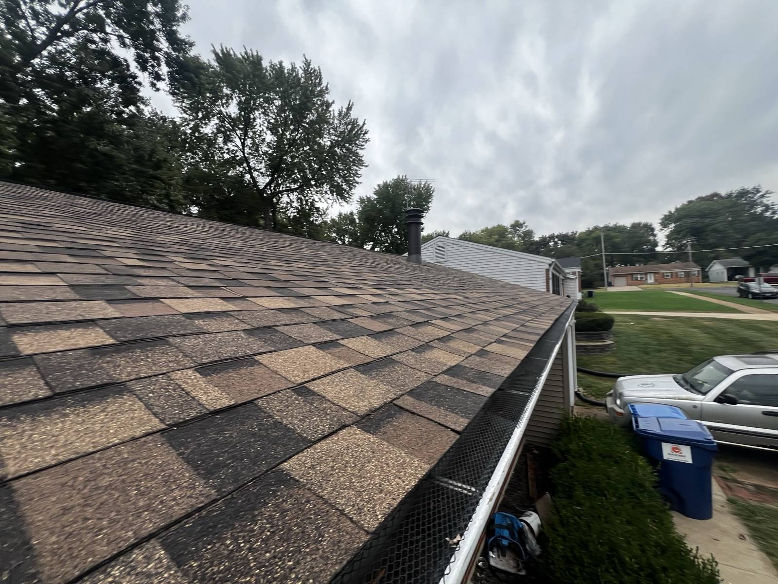 A weathered brown shingle roof photographed along the eave, with a mesh gutter guard and an ordinary suburban street below